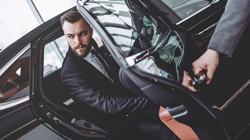 Jeune homme, cheveux en arrière, en costume sortant d'un véhicule de luxe avec chauffeur