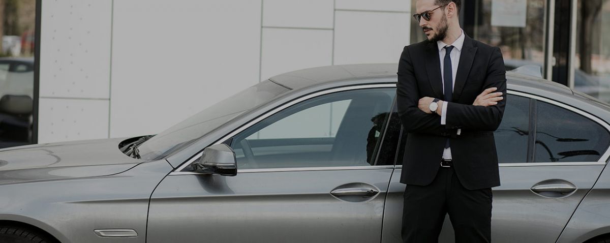 Dark-haired man in a suit waiting with his arms crossed, leaning against a grey car.
